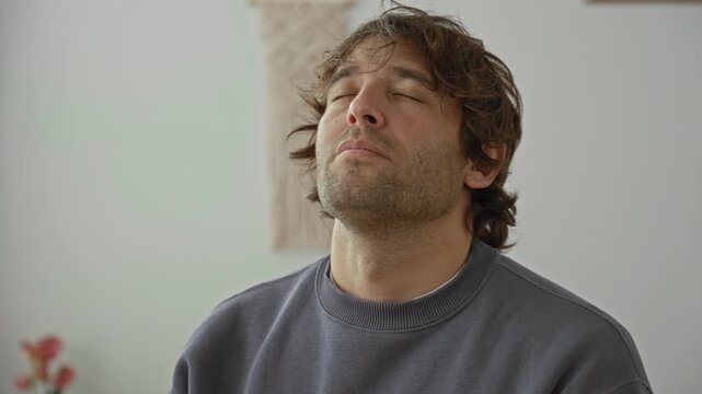 Man closes eyes and tilts head back showing exposed neck in studio, seated on a chair near macrame wall hanging and potted plant; calm mindfulness.