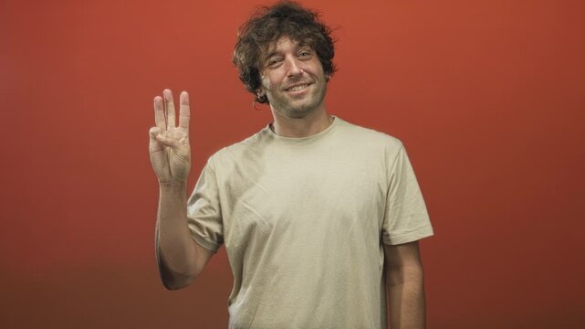 Young man holds up three fingers in a studio red backdrop, smiling as he shifts finger counts across frames and gestures to camera; counting playful confidence.