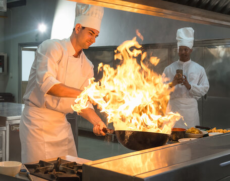 focused professional male chef in white uniform cooking with intense high flames and wok tossing in commercial restaurant kitchen, while coworker holds tablet in background.