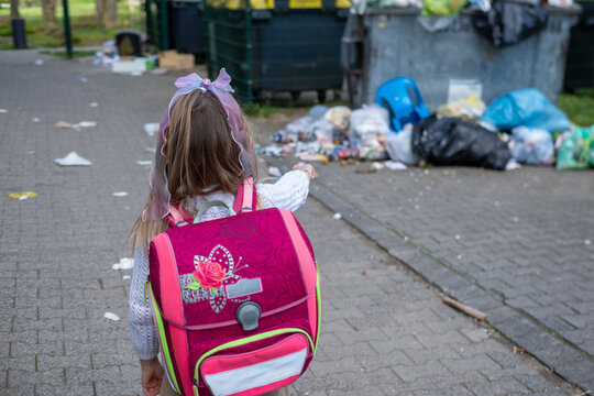 Little girl with pink school backpack pointing pile garbage near dumpsters, back view, environmental pollution concept, littering problem, waste management issue, urban trash, social commentary