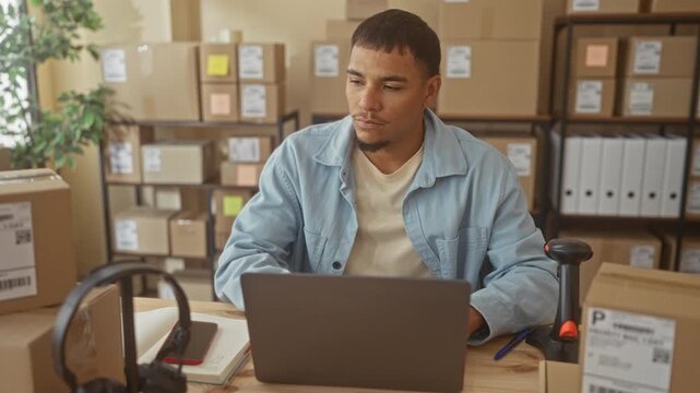 Man reaching hand to cardboard box at laptop on desk in studio surrounded by stacked shipping boxes and barcode scanner; determined entrepreneur hustle.