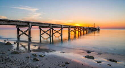 Fototapeta premium Long wooden pier extends over calm ocean waters during a serene sunset.