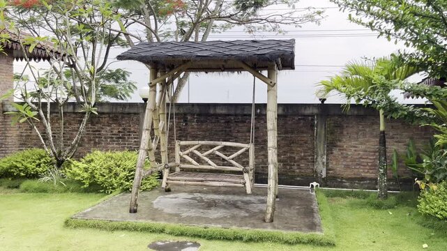 Rustic bamboo swing gazebo in a green garden courtyard with tropical plants and brick wall background.