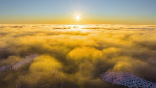 Aerial pan shot capturing a bright golden sun over a dense sea of clouds, revealing a vague snowy woodland through the misty cloud inversion and orange horizon.