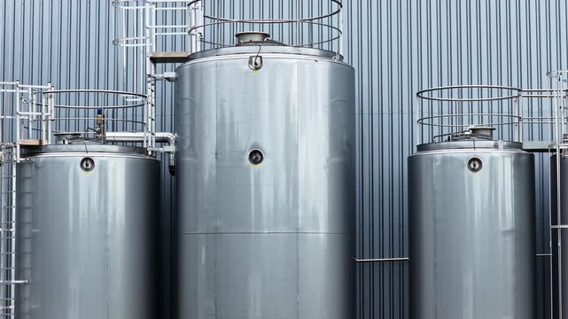 Drone rises along large stainless steel cylindrical storage tanks with safety railings, access hatches and pipework against corrugated metal industrial building facade