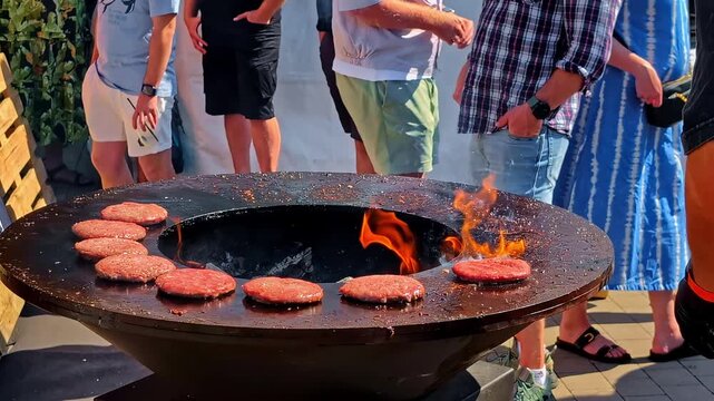 People cooking burgers on large outdoor circular grill with open flame during social gathering, friends walk around