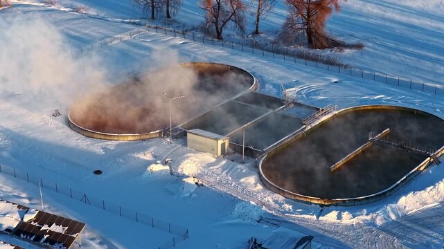 Drone flyover of steaming circular wastewater treatment clarifier tanks surrounded by snow at winter sunset in Latvia with solar panels nearby