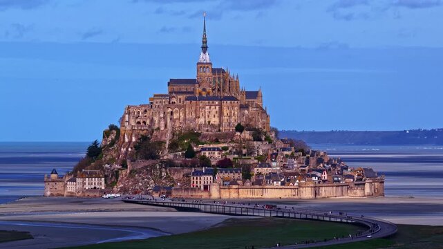 Cinematic aerial timelapse at blue hour showing Mont Saint Michel abbey glowing above tidal flats while streams of tourists move along the curving access bridge in Normandy France