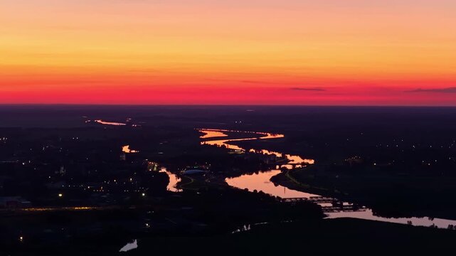 Aerial view of Lielupe River snaking through dark Jelgava city at sunrise, with blazing orange light dramatically reflected on winding water surface against shadowy landscape