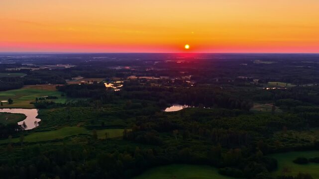 Drone aerial at sunset over patchwork fields, forests and small lakes, with a glowing orange sky and sun on the horizon. Peaceful rural panorama with cinematic color.