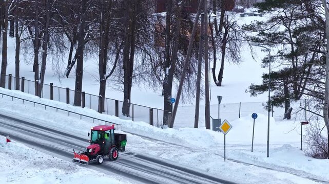 Small red tractor with front plow blade and rear spreader clearing snow on city road surrounded by trees and snowbanks in Latvia