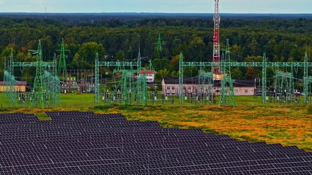 Aerial shot reveals solar panel array in foreground alongside large green high-voltage electrical substation with transmission towers, orange wildflower meadow and dense forest backdrop