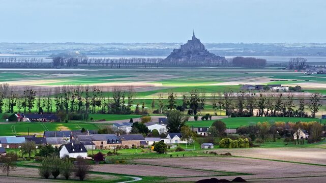 Aerial timelapse over traditional Norman hamlet and patchwork fields with iconic Mont Saint-Michel abbey silhouetted on distant tidal bay horizon in Normandy, France