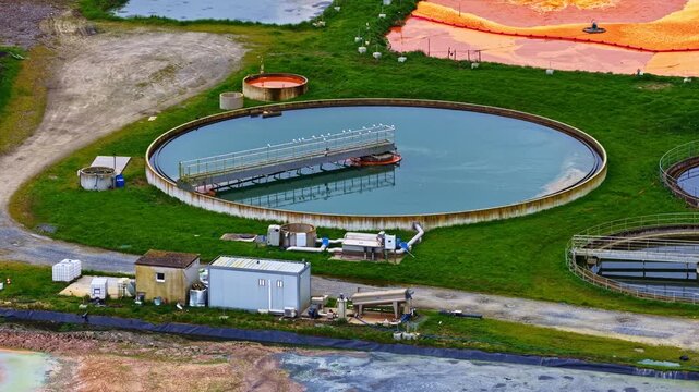 Drone parallax move reveals circular clarifier settling tank and vivid orange sludge oxidation beds with aeration nozzles at industrial wastewater treatment facility surrounded by green embankments