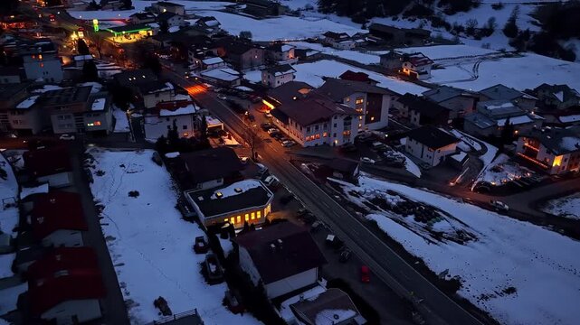 Drone orbits above snow-covered Nauders alpine village at night, revealing glowing street lights, illuminated buildings and snowy rooftops against dark winter landscape in Austria
