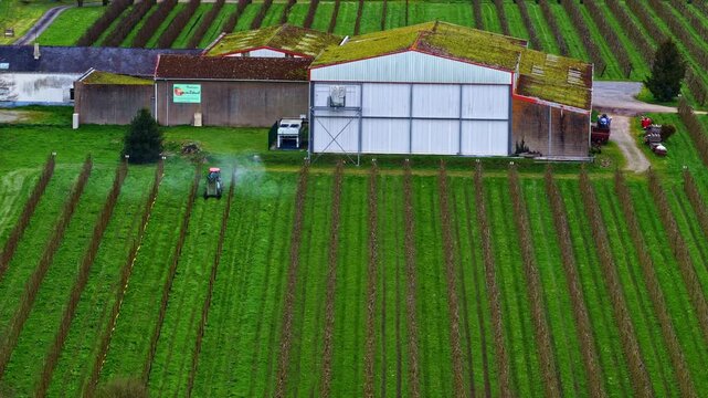 Drone captures red tractor spraying pesticide along neat green orchard rows near farm warehouse and storage sheds in Brittany, France