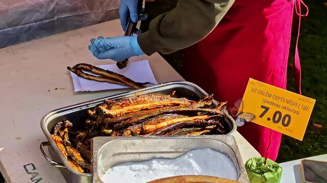 Latvian street market vendor serving charcoal-roasted lamprey (neģis) from a tray with tongs on display at an outdoor food stall.