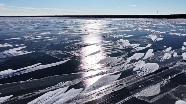 Low drone shot skimming frozen lake surface with black ice, scattered ice fragments and brilliant sun reflection stretching to horizon in Latvia