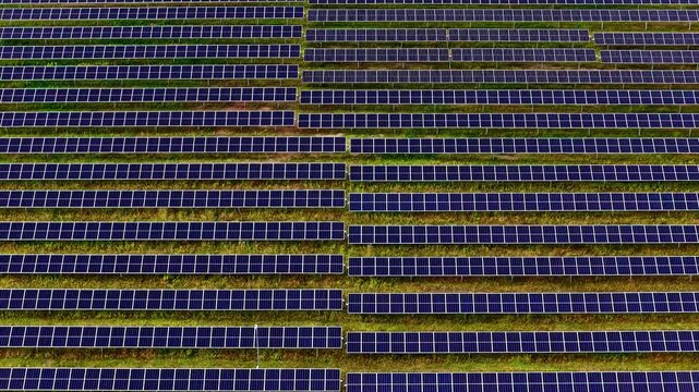 Drone flies sideways parallel to hundreds of photovoltaic solar panel rows creating mesmerizing geometric grid pattern with green grass strips between rows filling entire frame