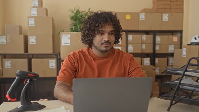 Man typing on laptop with barcode scanner and cardboard box, face visible and focused in building; determination entrepreneurship.