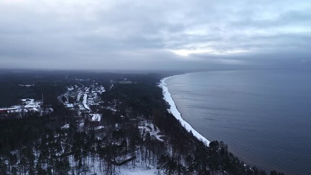 Drone aerial of a snowy forested coastline with a long curving beach and calm dark sea under overcast winter clouds, creating a moody Baltic shoreline panorama.