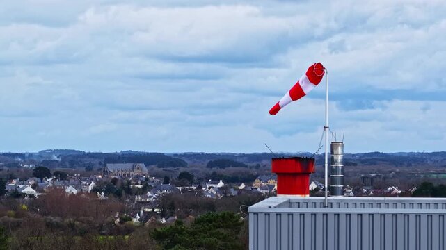 Windsock above an industrial rooftop with a distant town skyline under heavy clouds, capturing wind direction, urban industry, and changing weather conditions.