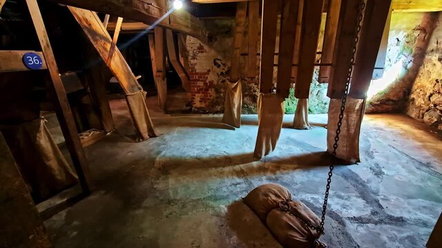 Old mill interior with hanging fabric chutes and a chained sandbag counterweight on a dusty floor. Rustic industrial heritage scene with moody light and texture.
