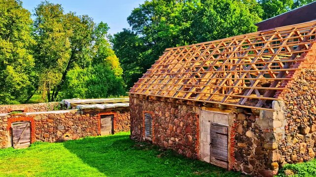 Stone outbuilding under renovation with exposed wooden roof frame and old doors, surrounded by green lawn and trees on a sunny day. Rustic heritage architecture restoration scene.