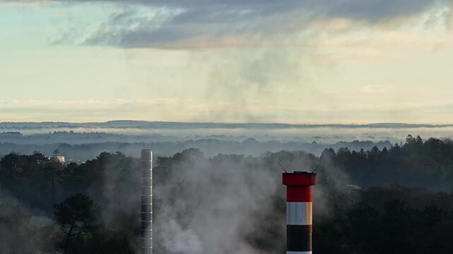 Industrial smokestacks from data center cloud processing server releasing steam above forest landscape under cloudy sky, aerial