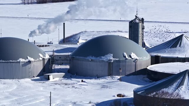 Aerial close-up of a snow-covered biogas plant with domed digesters, silos, steam plumes, and birds, capturing winter energy infrastructure and industrial operations.