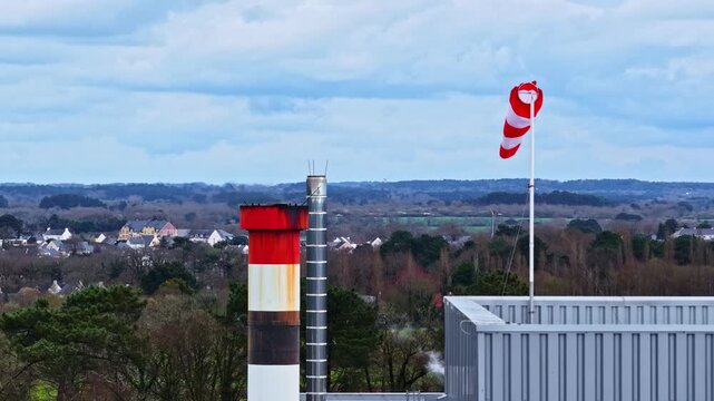 Windsock and industrial chimney with sno smoke rising above buildings and distant town landscape under cloudy sky, aerial