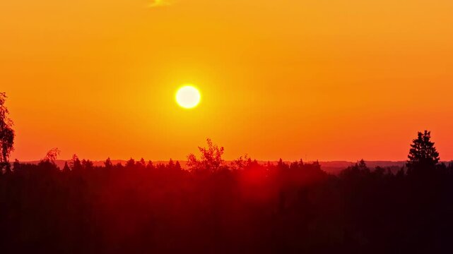 Drone slowly descends toward a dark forest silhouette at vivid orange sunset, glowing sun over distant horizon with warm atmospheric haze.