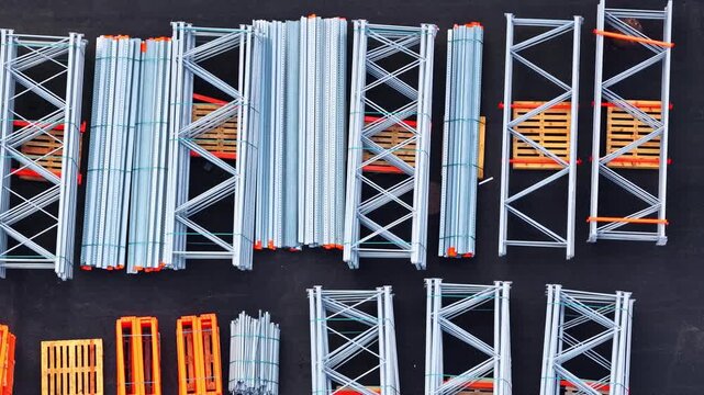 Drone flies sideways top-down view of galvanized steel pallet racking frames, orange brackets and wooden pallets neatly arranged on dark asphalt in industrial yard