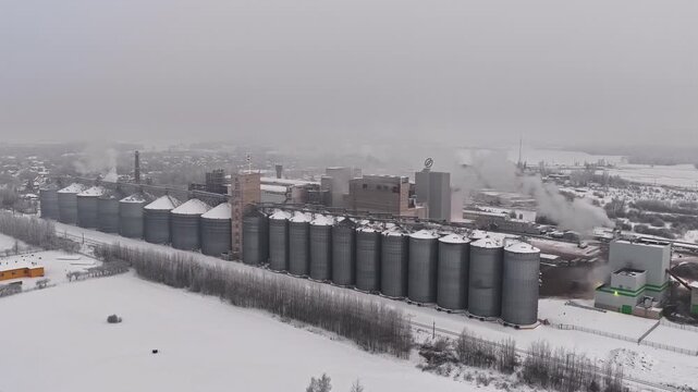 Aerial drone reveal of large grain silo complex and processing plant covered in snow with steam rising from chimneys in winter at Jelgava, Latvia