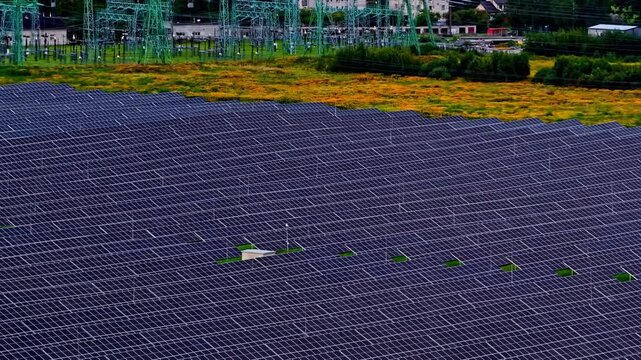 Drone tilt-up shot reveals vast solar panel array next to high-voltage power substation with green transmission towers, meadow, and forest