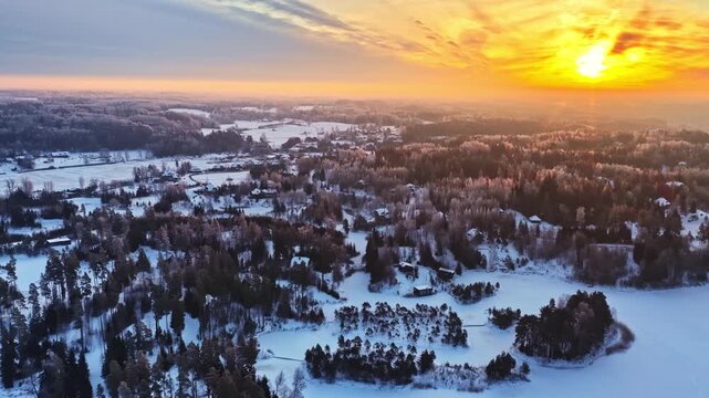 Aerial drone slowly flying over snowy countryside landscape with forests and scattered houses at sunrise.