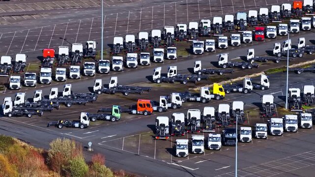 Rows of truck chassis parked in large logistics yard with organized parking lines and industrial asphalt lot, aerial telephoto orbit around tractors