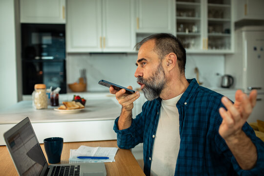 Bearded man using voice assistant on smartphone while working in home kitchen
