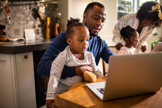 Father helping toddler use laptop at kitchen table at home