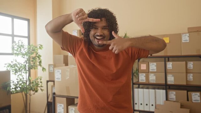 Man smiling amid stacked cardboard boxes in an office building while framing parcels with his hands; entrepreneurial confidence.