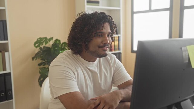 Man covering mouth with hands at computer in office building, white t-shirt, curly hair, bookshelf and plant visible, sticky note on monitor; joy celebration.