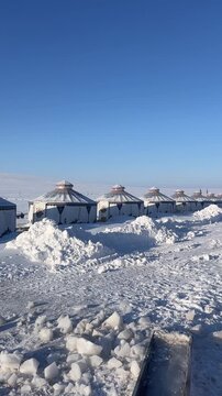 A peaceful vertical view of traditional Mongolian yurts covered in deep snow on a vast frozen steppe. Smoke gently rises from a chimney.
