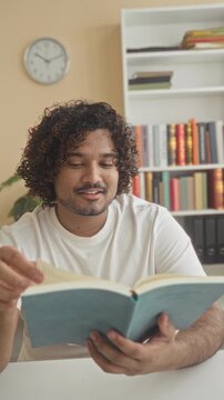 Man reading a book with hand on chin, smiling while turning a page at a table with bookshelf and clock in a building; serenity focus learning.