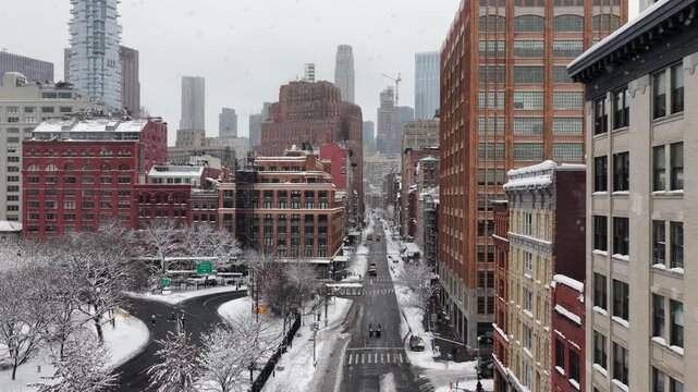 Aerial wide shot of snowy New York City streets lined with classic brick buildings, as light snow flurries fall over quiet traffic and curved roads, revealing dense urban architecture. Wide shot.