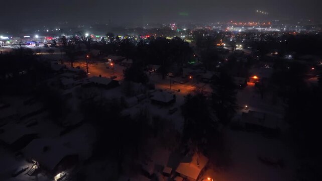 Aerial night flyover of snow-covered suburban neighborhood in America, with warm streetlights glowing over quiet houses and roads. Calm winter atmosphere under dark sky. Downtown lights in background.