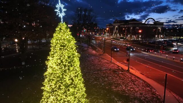 Aerial view of illuminated Christmas tree with star during snowfall at dusk in Hershey, Pennsylvania with passing traffic and festive lights. Warm holiday atmosphere in small town in United States.