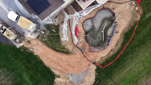 Top-down aerial view of custom gunite swimming pool construction in suburban backyard. Drone shot showing concrete framework, excavation and modern house with solar panels on sunny day.