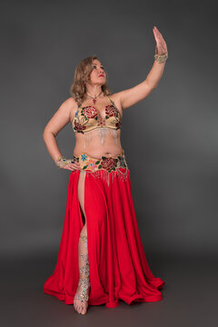 Authentic mid aged adult woman belly dancer and fashion model donning in dancing stage costume - bra top and scarlet long skirt with slit moves barefoot in full length against gray background