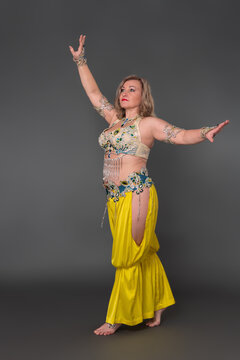 Blonde woman belly dancer dressing in dancing stage costume - bra top and yellow harem trousers and she is not wearing any footwear standing in full length against gray background