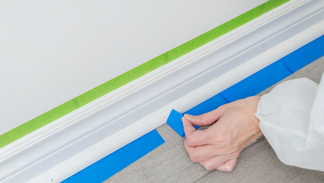 Person applying blue painter's tape along baseboard in preparation for painting, with green tape visible on the wall above